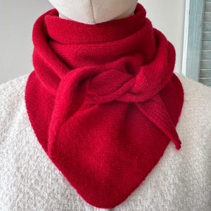 May include: A vibrant red knitted scarf featuring a triangular shape and a textured stitch pattern. The scarf is wrapped around the neck with a knotted detail at the front, creating a layered appearance with pointed ends that drape downward.