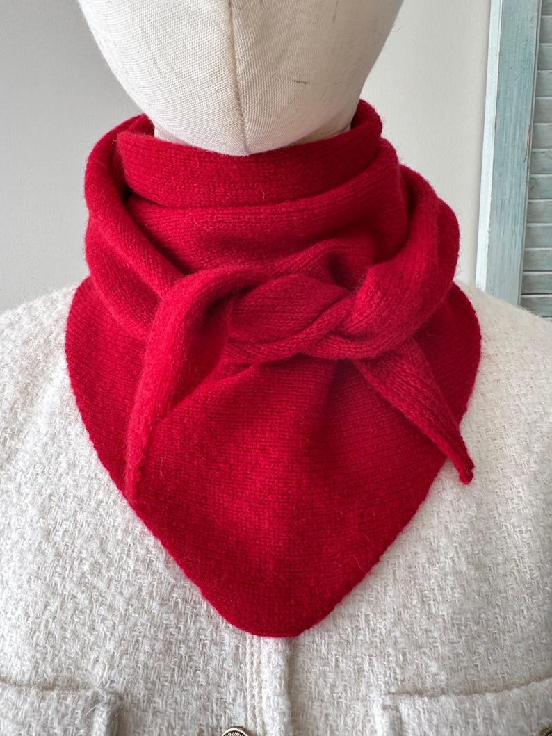 May include: A vibrant red knitted scarf featuring a triangular shape and a textured stitch pattern. The scarf is wrapped around the neck with a knotted detail at the front, creating a layered appearance with pointed ends that drape downward.