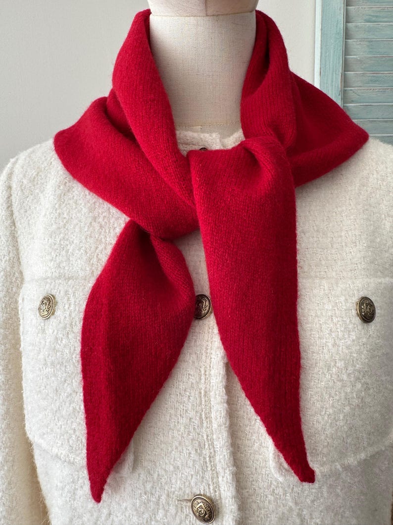 May include: A bright red knit scarf tied around the neck, worn over a white textured jacket featuring gold-toned buttons. The scarf has a soft, matte appearance and is draped with two pointed ends hanging down the front of the white garment.
