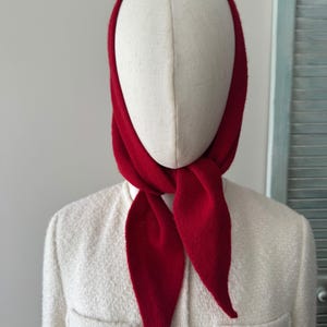 May include: A bright red knit headscarf tied under the chin, worn over a cream-colored textured jacket with gold-toned buttons. The headscarf features a soft, matte finish and a triangular shape that drapes over the head and neck area.