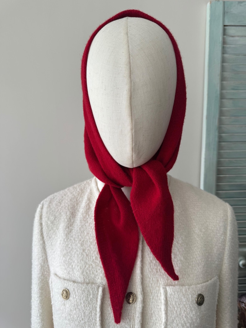 May include: A bright red knit headscarf tied under the chin, worn over a cream-colored textured jacket with gold-toned buttons. The headscarf features a soft, matte finish and a triangular shape that drapes over the head and neck area.