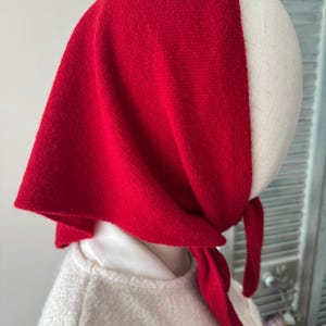 May include: A vibrant red triangular headscarf tied securely under the chin. The fabric has a soft, knit texture and drapes over the head, covering the top and sides. The scarf is positioned on a mannequin head, showing a simple knotted closure.
