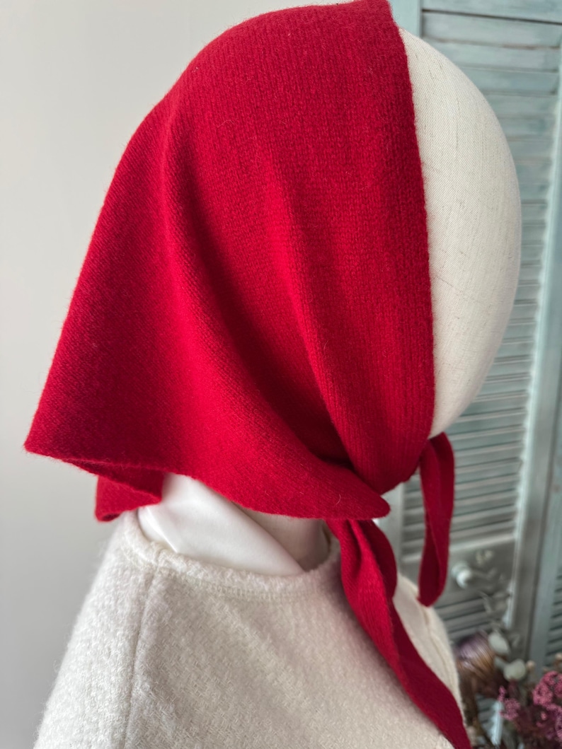 May include: A vibrant red triangular headscarf tied securely under the chin. The fabric has a soft, knit texture and drapes over the head, covering the top and sides. The scarf is positioned on a mannequin head, showing a simple knotted closure.