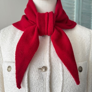 May include: A bright red knitted triangular scarf tied in a knot around the neck, worn over a white textured jacket featuring gold-toned buttons. The scarf has a soft, ribbed knit texture and pointed ends that drape down the front of the jacket.