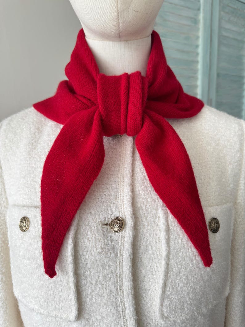 May include: A bright red knitted triangular scarf tied in a knot around the neck, worn over a white textured jacket featuring gold-toned buttons. The scarf has a soft, ribbed knit texture and pointed ends that drape down the front of the jacket.