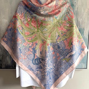 May include: A square silk scarf featuring a detailed jungle scene with green palm trees, blue leopard figures, and intricate floral patterns in shades of pink, blue, and green. The scarf has a solid light pink border and is draped over a white shirt.