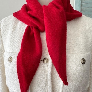 May include: A bright red knit scarf tied around the neck, worn over a cream-colored textured jacket. The jacket features two chest pockets and is fastened with round, metallic gold-toned buttons that have a detailed embossed design.