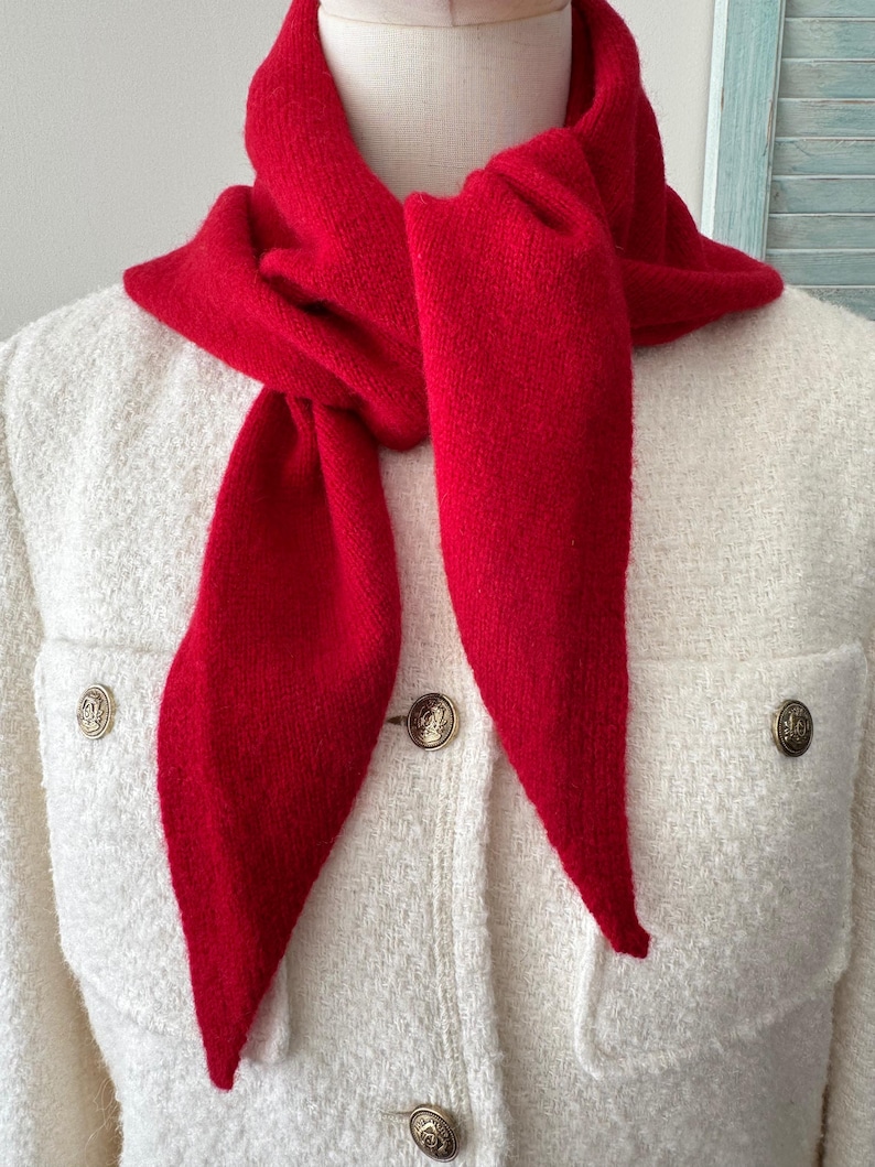 May include: A bright red knit scarf tied around the neck, worn over a cream-colored textured jacket. The jacket features two chest pockets and is fastened with round, metallic gold-toned buttons that have a detailed embossed design.