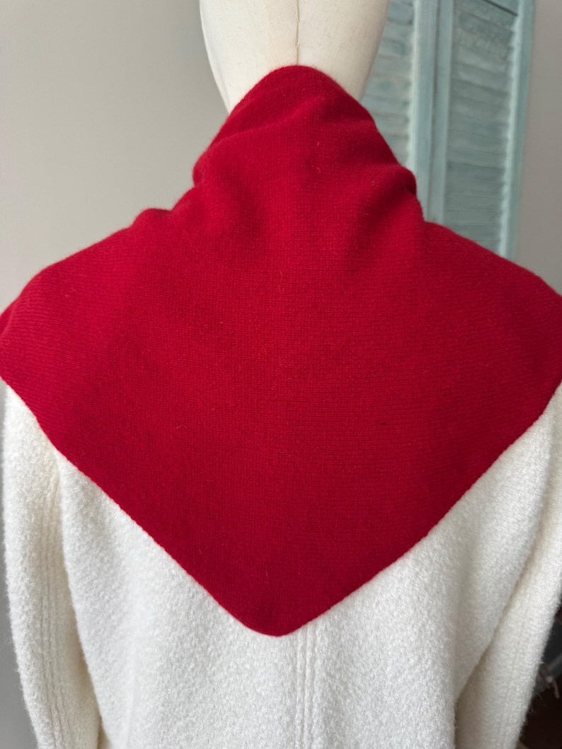 May include: A white knit sweater featuring a prominent, solid red triangular-shaped panel across the upper back and shoulders. The garment has a high neckline and a textured, woven appearance, displayed on a mannequin to show the back view.