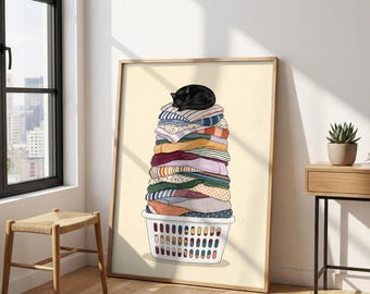 Op de afbeelding: Een ingelijste kunstdruk met een witte wasmand die overloopt van een hoge, wankele stapel gevouwen kledingstukken in diverse kleuren en patronen, met bovenop een slapende zwarte kat tegen een licht crèmekleurige achtergrond.
