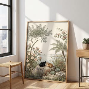 May include: A framed art print showing a tranquil landscape with two cats. The artwork features a black cat and a ginger cat on a rock surrounded by greenery, flowers, and a coastal view. The frame is a light wood colour.