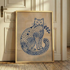 Op de afbeelding: Een ingelijste kunstprint met een blauwe inktillustratie van een zittende kat op een beige achtergrond. De kat is versierd met ingewikkelde stippen en wervelende patronen en houdt een takje met bessen in de bek in een houten lijst.