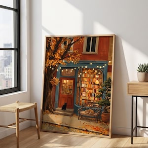 May include: A framed art print depicting a bookshop exterior with a red door, a large window filled with books, string lights, a black cat on the steps, and a cat resting on a bench. The scene features an autumn tree with orange leaves.