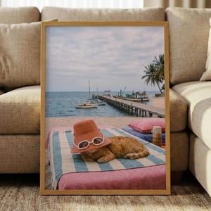 May include: A framed print featuring a ginger cat wearing a straw hat and sunglasses, lounging on a beach towel. The background shows a pier, boats, and a tropical beach scene with palm trees.