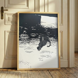 May include: A framed black and white art print depicting a cat's reflection in water with concentric ripples. The artwork is in a light wood frame and leans against a white door and wooden floor.
