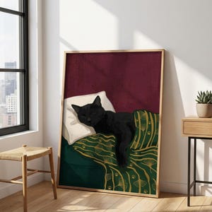 May include: A framed art print depicts a black cat sleeping on a pillow and under a green blanket with a gold leaf pattern. The background is a deep burgundy color. The artwork is displayed in a room with a window and a wooden stool.