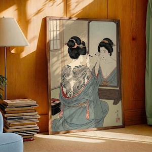 以下が含まれることがあります： 鏡の前で化粧をする着物姿の女性を描いた額装されたアートワーク。女性の背中には大きな龍のタトゥーがあります。このアートワークは、柔らかい色と繊細なラインで、伝統的な日本のスタイルです。
