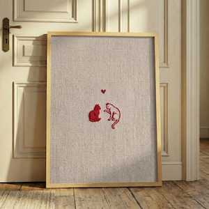 May include: A framed wall art piece featuring a light beige textured fabric background. Centered on the fabric are two embroidered red cat silhouettes facing each other with a small red embroidered heart positioned above them.