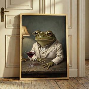 May include: A framed wall art print depicting a green frog wearing round gold-rimmed glasses and a beige suit jacket. The frog sits at a wooden table with a martini glass filled with a dark red liquid. A lamp base is visible on the left side.