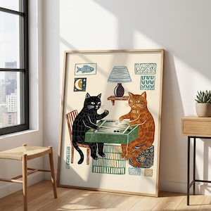 May include: A framed art print featuring two cats playing backgammon. One cat is black, the other is orange. The print has a light beige background with decorative elements like a fish, moon, and lamp.