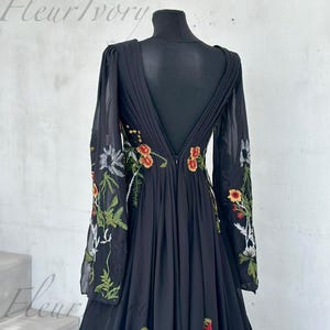 May include: Black floor-length gown with floral embroidery. The dress features long, sheer sleeves and a deep V-back. The floral embroidery includes red, orange, and green accents. The dress is displayed on a black mannequin.