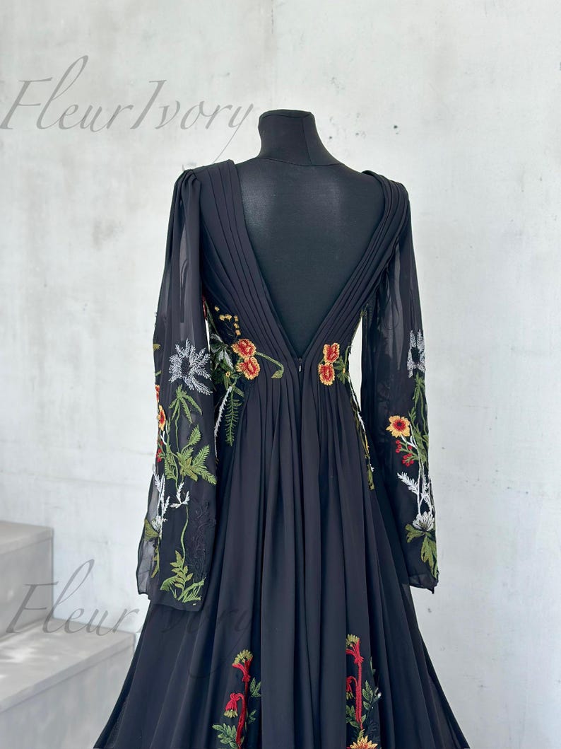 May include: Black floor-length gown with floral embroidery. The dress features long, sheer sleeves and a deep V-back. The floral embroidery includes red, orange, and green accents. The dress is displayed on a black mannequin.