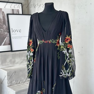 May include: Black chiffon dress with a deep V-neck and long, sheer sleeves. The dress features intricate floral embroidery in red, orange, and white along the sleeves and waist. The dress is displayed on a black mannequin.