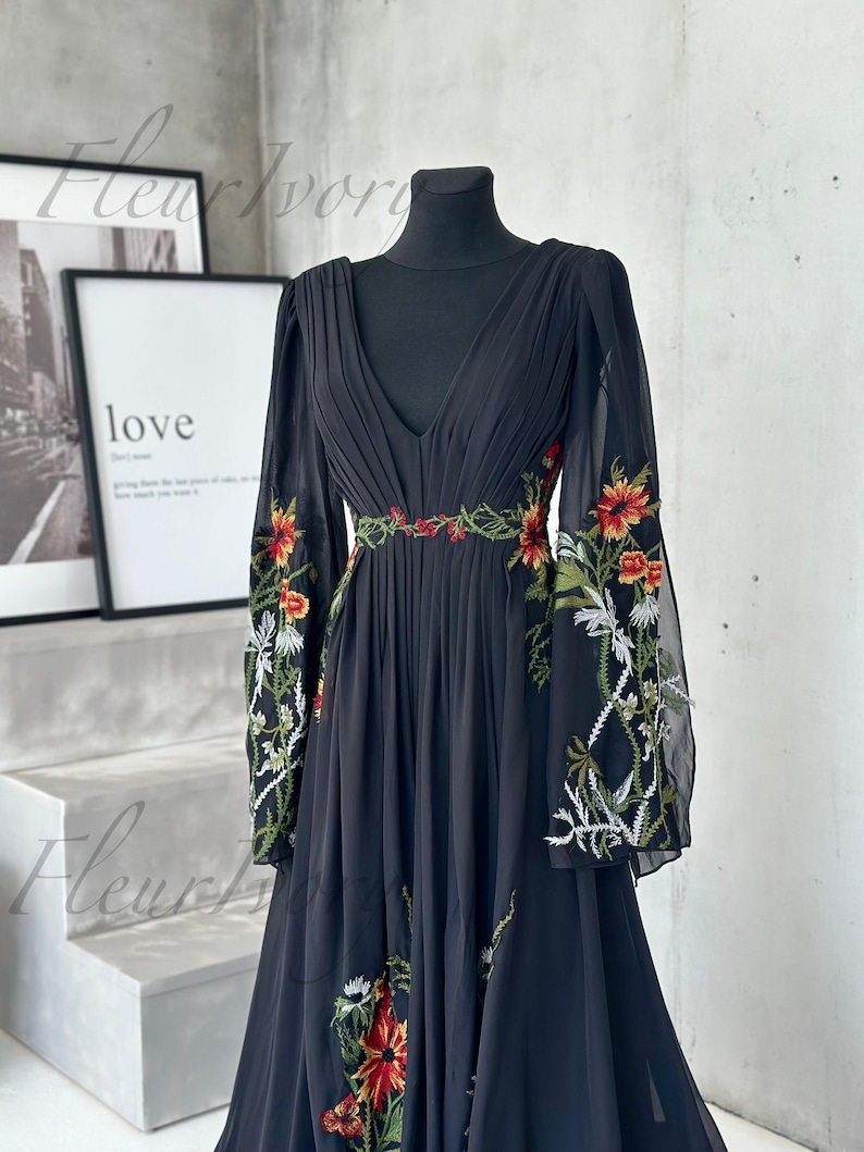 May include: Black chiffon dress with a deep V-neck and long, sheer sleeves. The dress features intricate floral embroidery in red, orange, and white along the sleeves and waist. The dress is displayed on a black mannequin.