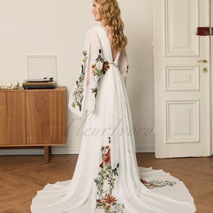 May include: A white wedding dress with floral embroidery. The dress features long, flowing sleeves with embroidered flowers and a deep V-back. The floral embroidery includes red, orange, and green accents. The dress has a long train and is made of a lightweight fabric.