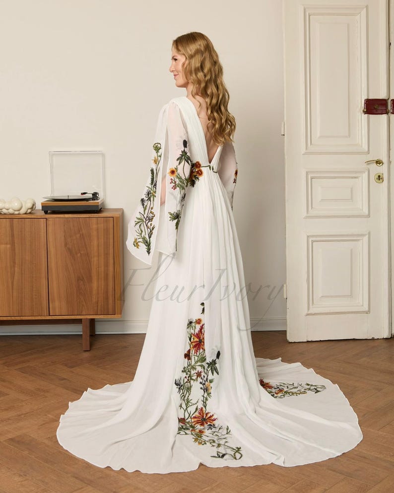 May include: A white wedding dress with floral embroidery. The dress features long, flowing sleeves with embroidered flowers and a deep V-back. The floral embroidery includes red, orange, and green accents. The dress has a long train and is made of a lightweight fabric.
