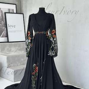 May include: Black floor-length dress with long sleeves and a V-neck. The dress features floral embroidery in red, orange, and white along the sleeves and waist. The dress is displayed on a black mannequin.