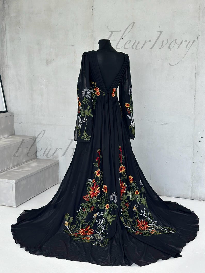May include: A black floor-length gown with long sleeves and a plunging neckline. The dress features intricate floral embroidery in shades of red, orange, green, and white. The dress is displayed on a black mannequin.