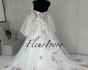 May include: A white off-the-shoulder gown featuring sheer puff sleeves and a sweetheart neckline. The dress is adorned with colorful floral embroidery in shades of purple, yellow, and orange. The text FleurIvory is centered on the skirt.