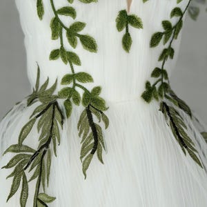 May include: A white dress featuring a deep V-neckline and intricate green leaf embroidery. The embroidery extends from the bodice down to the waistline, showcasing detailed stitching of foliage patterns on a sheer, layered tulle fabric.