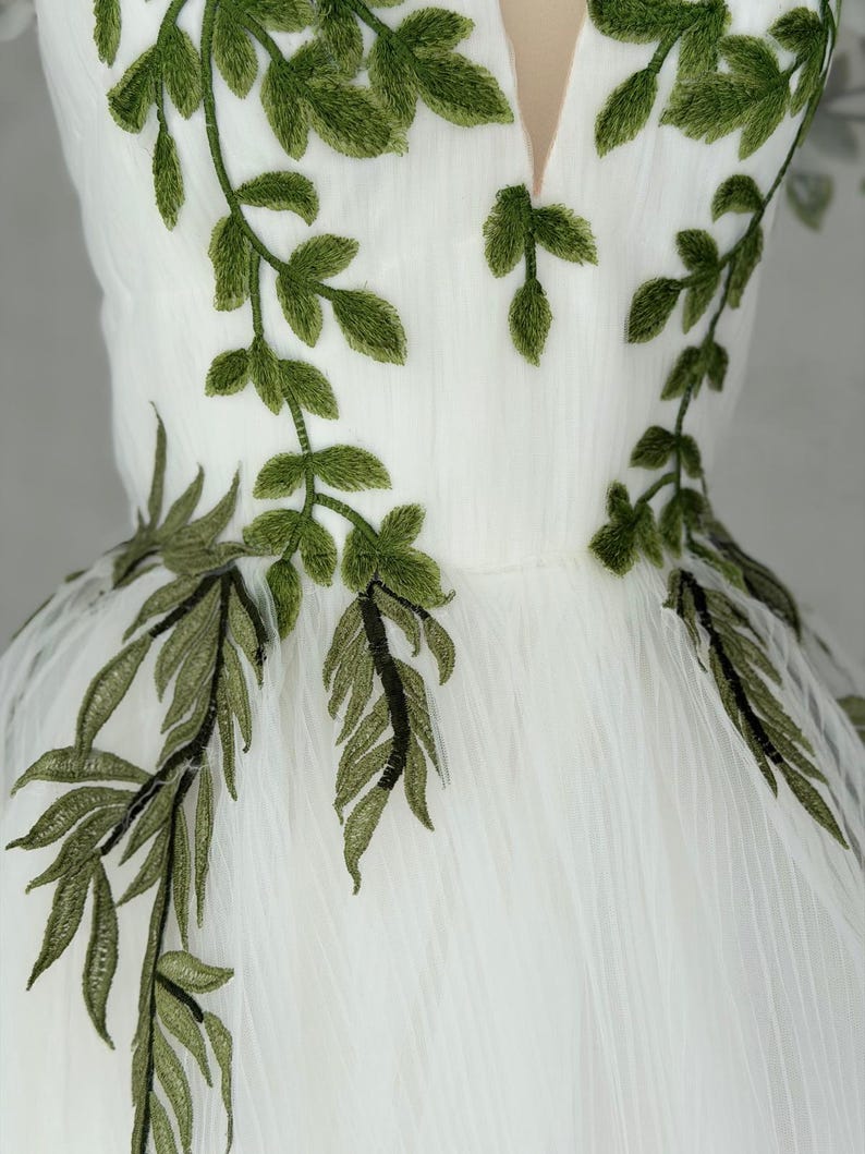 May include: A white dress featuring a deep V-neckline and intricate green leaf embroidery. The embroidery extends from the bodice down to the waistline, showcasing detailed stitching of foliage patterns on a sheer, layered tulle fabric.
