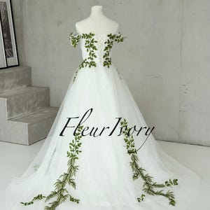 May include: A floor-length white tulle gown featuring off-the-shoulder sleeves and a lace-up back closure. The dress is decorated with green embroidered vine and leaf patterns that extend from the bodice down to the hem. The text Fleur Ivory is visible.