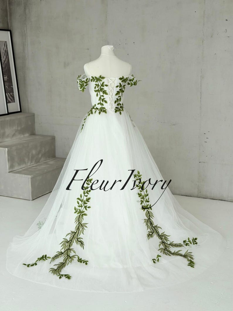 May include: A floor-length white tulle gown featuring off-the-shoulder sleeves and a lace-up back closure. The dress is decorated with green embroidered vine and leaf patterns that extend from the bodice down to the hem. The text Fleur Ivory is visible.