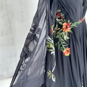 May include: Black chiffon dress with floral embroidery. The dress features a V-neckline and flowing sleeves. The embroidery includes red, yellow, and green flowers and leaves. The dress is displayed on a mannequin.