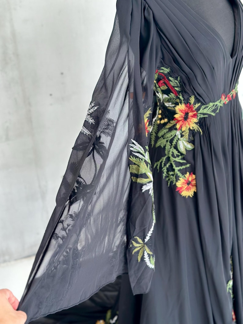 May include: Black chiffon dress with floral embroidery. The dress features a V-neckline and flowing sleeves. The embroidery includes red, yellow, and green flowers and leaves. The dress is displayed on a mannequin.