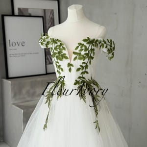 May include: A white off-the-shoulder gown featuring a deep V-neckline and sheer straps. The bodice is adorned with green leaf embroidery that extends from the neckline to the waist. The skirt is made of layered tulle fabric.