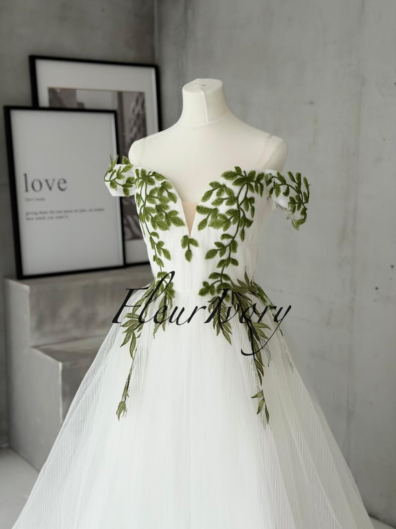 May include: A white off-the-shoulder gown featuring a deep V-neckline and sheer straps. The bodice is adorned with green leaf embroidery that extends from the neckline to the waist. The skirt is made of layered tulle fabric.