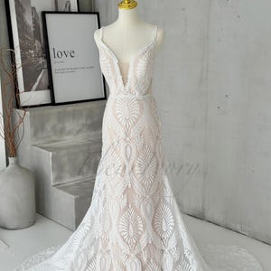 May include: A white wedding dress with a plunging neckline and intricate lace detailing. The dress features a fitted bodice that flares into a mermaid silhouette with a long train. The dress is displayed on a mannequin.
