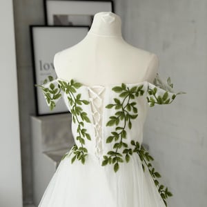 May include: A white off-the-shoulder dress featuring green leaf embroidery along the bodice and sleeves. The back of the garment includes a white lace-up corset closure with matching white ribbon, set against a sheer, layered tulle skirt.