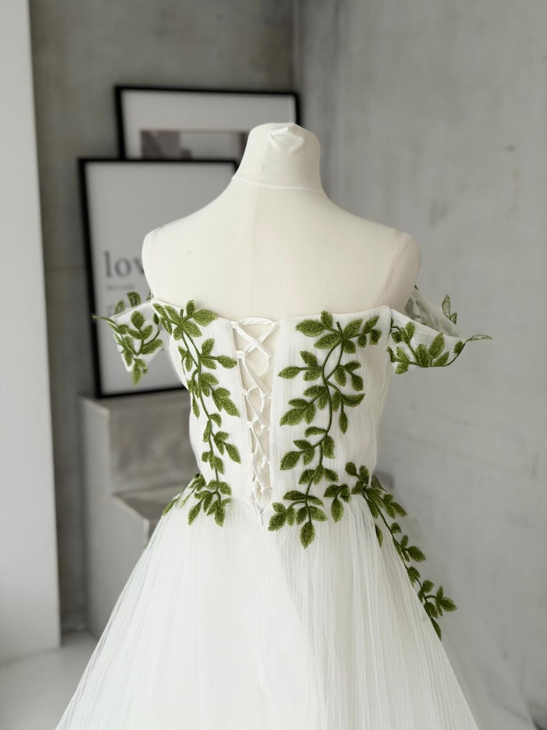 May include: A white off-the-shoulder dress featuring green leaf embroidery along the bodice and sleeves. The back of the garment includes a white lace-up corset closure with matching white ribbon, set against a sheer, layered tulle skirt.