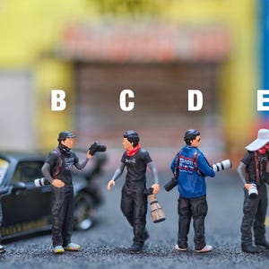 May include: Miniature figurines of photographers in various poses, some holding cameras and lenses. One figure wears a red shirt, while others sport black shirts and jackets. A black sports car is in the background.