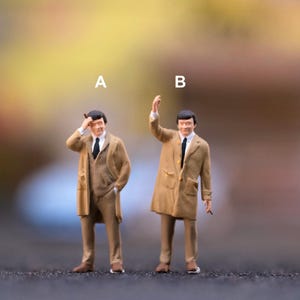 Può includere: Figurine in miniatura di due uomini in abiti e cappotti beige. Una figura si tocca la testa, l'altra alza la mano. Entrambe le figure indossano cravatte nere e scarpe marroni. Lo sfondo è sfocato con toni gialli e marroni.