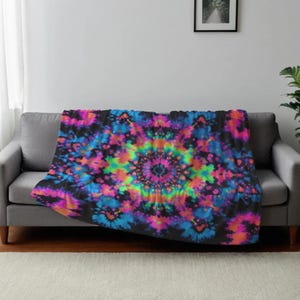Może przedstawiać: Kolorowy koc typu tie-dye z żywym wzorem mandali. Koc ma czarne tło z okrągłym wzorem w odcieniach różu, błękitu, zieleni i pomarańczy. Jest narzucony na szarą sofę.