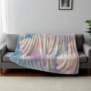 May include: A soft, plush throw blanket with a watercolor-style design featuring clouds in shades of blue, pink, and yellow. The blanket is draped over a gray couch.