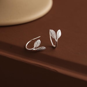 Puede incluir: Un par de puños de oreja plateados con forma de hoja. Cada puño presenta un delicado diseño de hoja con detalles intrincados. Los pendientes se exhiben sobre una superficie marrón, mostrando su estética elegante e inspirada en la naturaleza.
