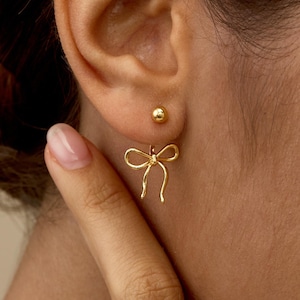 Könnte beinhalten: Ein Paar goldfarbene Ohrringe in Schleifenform. Ein Ohrring ist eine kleine goldene Kugel, der andere ein filigranes Schleifendesign. Die Ohrringe werden an einem Ohr getragen.
